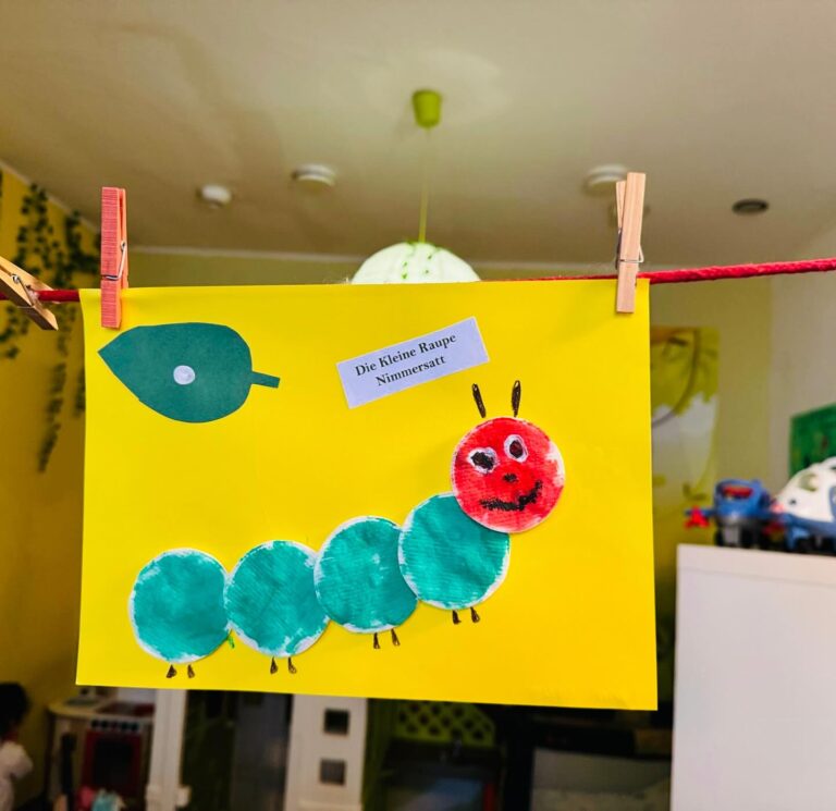Selbstgebasteltes Bild einer kleinen Raupe Nimmersatt aus buntem Papier, aufgehängt an einer roten Schnur mit Wäscheklammern.
